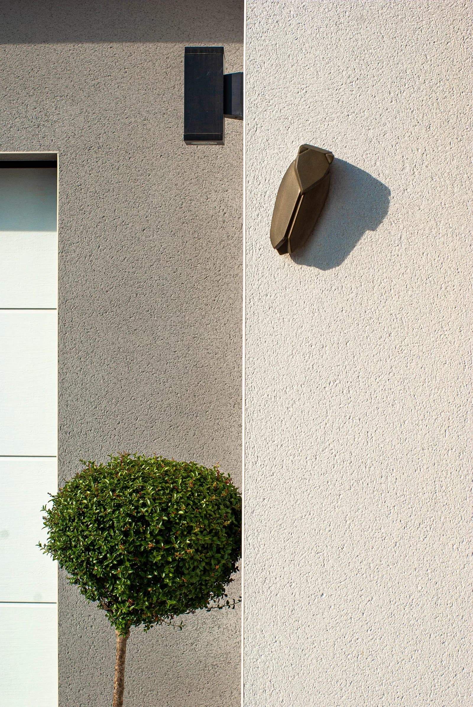 Détail d’un enduit clair, d’éclairages extérieurs et d’une entrée plantée.