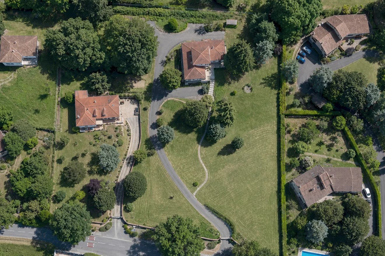 Vue aérienne d’une grande parcelle enherbée traversée par une voie d’accès et entourée d’habitations.